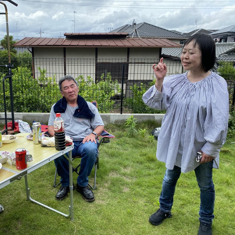 庭で、女性が身ぶりをまじえて話し、椅子に座った男性が聞いている写真